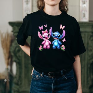 May include: Black t-shirt featuring a colorful graphic of two cartoon characters, one pink and one blue, with butterflies and flowers. The characters are holding hands. The shirt is made of a soft material.