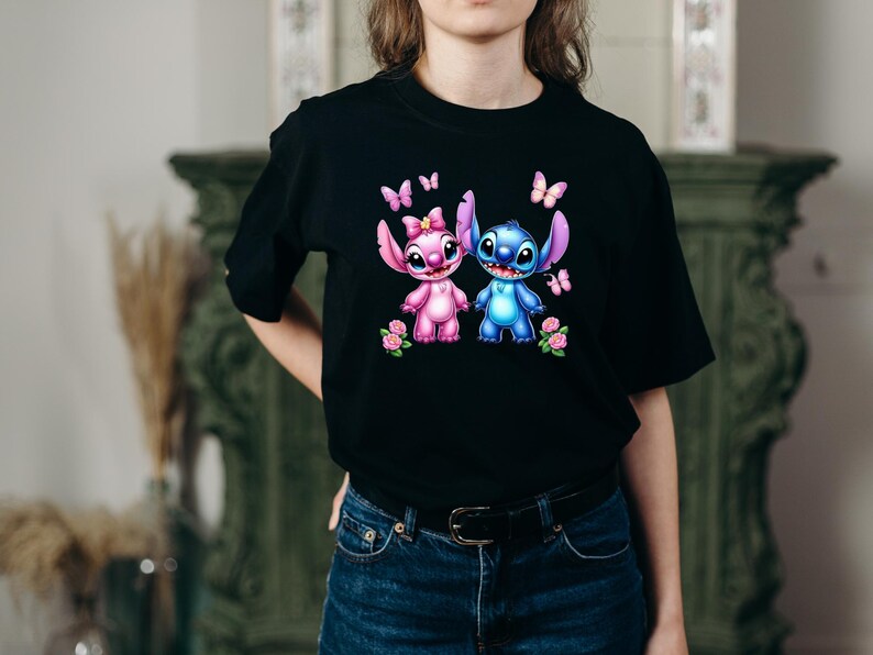 May include: Black t-shirt featuring a colorful graphic of two cartoon characters, one pink and one blue, with butterflies and flowers. The characters are holding hands. The shirt is made of a soft material.