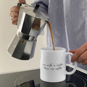 Può includere: Una caffettiera italiana argentata versa il caffè in una tazza bianca. La tazza ha il testo scritto a mano "my wife is hotter than my coffee". La persona indossa una camicia a righe blu e bianche.