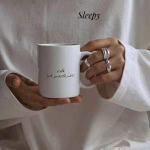 Può includere: Tazza in ceramica bianca con la scritta "cute but overthinker" in corsivo nero. La tazza è tenuta davanti a una maglietta bianca a maniche lunghe con la parola "Sleepy" stampata. Diversi anelli d'argento adornano la mano.