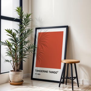 May include: A framed art print featuring a large block of orange with the text "Tangerine Tango" and a color code. A tall green plant in a beige pot sits next to the print. A small wooden stool with black legs is also in the image.