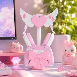 Könnte beinhalten: Ein rosa-weißer Gaming-Headset- und Controller-Ständer mit einem zentralen Herz-Emblem mit weißen Flügeln, einer großen rosa Schleife am Controller-Halter und einem stabilen Sockel zur organisierten Aufbewahrung von Gaming-Zubehör.