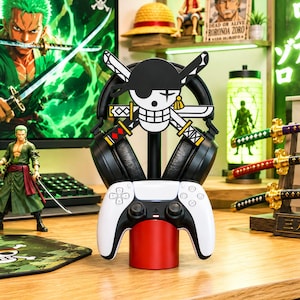 May include: A black gaming headset stand featuring a skull and crossbones emblem with swords, supporting a pair of black over-ear headphones and a white and black wireless game controller on a cylindrical red base, designed for gaming desk organisation.