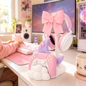 Könnte beinhalten: Ein weißer, wolkenförmiger Gaming-Zubehörständer mit einer großen rosa Schleife an der Spitze, einem lavendelfarbenen Haifischflossen-Akzent mit zwei kleinen Sternsymbolen und einem rosa-weißen Gamecontroller auf der Basis mit weißem Kopfhörer.