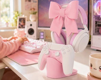 Pode incluir: Um suporte para acessórios de jogos em rosa claro com um grande laço decorativo no topo. O suporte segura um par de auscultadores brancos e rosa claro e um comando de jogo sem fios branco e rosa claro sobre uma base rosa a condizer.
