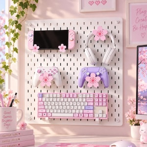 Puede incluir: Un organizador de tablero perforado blanco que muestra una consola de juegos portátil, auriculares blancos, dos mandos y un teclado mecánico, decorados con detalles de flores de cerezo rosas. Incluye una taza con texto y libros apilados.