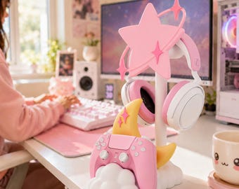 Pode incluir: Um suporte branco com uma estrela rosa no topo e uma lua crescente amarela na base, projetado para segurar um par de auscultadores brancos e rosa e um comando de jogo sem fios rosa, tudo assente numa base em forma de nuvem branca.