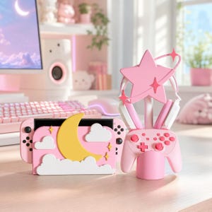 Puede incluir: Una base de carga para consola de juegos rosa con una luna creciente amarilla y nubes blancas, junto a un soporte para mando rosa a juego. El soporte sostiene un mando rosa y unos auriculares decorados con una gran estrella rosa.