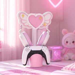 Op de afbeelding: Een gaming-opstelling met een wit en roze hartvormige koptelefoonstandaard met vleugels, een witte gamecontroller, een roze mechanisch toetsenbord, een roze muis en een monitor met een roze landschap en een digitale klok op 12:00.