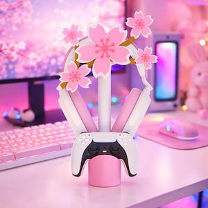 Op de afbeelding: Een wit en roze gaming headset standaard met een centrale verticale paal bekroond met decoratieve roze kersenbloesems. De standaard bevat een basis ontworpen om een wit en zwart spelbesturingsapparaat vast te houden, met de headset erbovenop.