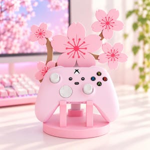 Op de afbeelding: Een lichtroze videogamecontroller rust op een ronde standaard met een decoratieve tak met vijf lichtroze kersenbloesems. De controller heeft witte thumbsticks, een wit richtingspad en standaard gekleurde actieknoppen.