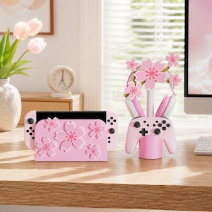 Può includere: Una base di ricarica per console di gioco rosa chiaro e un supporto per controller abbinato, entrambi decorati con motivi di fiori di ciliegio bianchi e rosa. Il supporto per controller presenta un montante verticale con sagome di fiori.