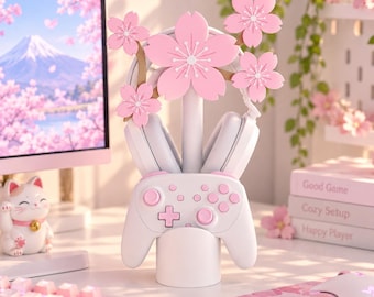 Pode incluir: Um suporte branco para comando de jogos e auscultadores com um poste vertical central decorado com cinco flores de cerejeira cor-de-rosa. O suporte foi concebido para segurar um comando de jogos sem fios branco na base e auscultadores acima.