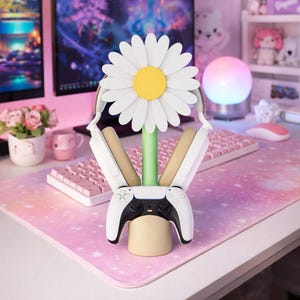 以下が含まれることがあります： 白と黄色のデイジー型のヘッドホンスタンド。緑色の茎とベージュのベースが付いています。白と黒のゲームコントローラーを保持しています。背景には、ピンクのキーボード、マウス、デスクマットが含まれています。