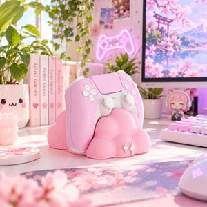 Pode incluir: Um suporte para comando de jogos em forma de nuvem rosa segurando um comando sem fios rosa e branco. O suporte apresenta um pequeno &iacute;cone de brilho branco na frente. O comando tem joysticks, bot&otilde;es e um painel t&aacute;til na cor branca.