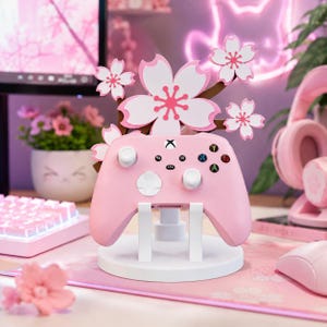 Puede incluir: Un mando de juego rosa claro descansa sobre un soporte circular blanco. Detrás del mando, una rama decorativa marrón presenta varias flores de cerezo blancas y rosas. El mando incluye botones estándar, joysticks y un logotipo de Xbox.