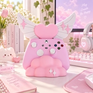 May include: A light pink gaming controller stand featuring a central heart shape with large white angel wings extending from the sides. The base is shaped like a cloud with two white star accents. The stand is designed to hold a standard game controller.