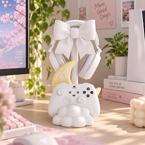 May include: A white gaming accessory stand featuring a large bow at the top, a crescent moon accent, and a cloud-shaped base. The stand holds a pair of white over-ear headphones and a white game controller with colored buttons and a small face icon.