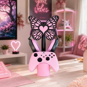 Könnte beinhalten: Ein rosa Gaming-Controller-Ständer mit einem schwarz-rosa schmetterlingsförmigen Aufsatz, der ein Herz in der Mitte zeigt. Der Ständer hält einen schwarz-rosa Kopfhörer und einen hellrosa kabellosen Gaming-Controller mit schwarzen Tasten.