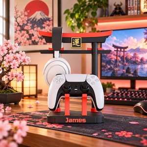 以下が含まれることがあります： 鳥居の形をした黒と赤のゲーミングコントローラーおよびヘッドセットスタンド。スタンドには金色の四角の中に幸運を表す日本語の漢字が記され、台座にはオレンジ色でJamesという名前が印刷されており、白いゲームコントローラーを保持しています。