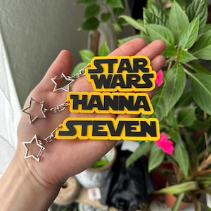 Op de afbeelding: Gele en zwarte Star Wars sleutelhangers met de namen Hanna en Steven. Elke sleutelhanger heeft een ster-vormige bedel die met een ketting is bevestigd. De sleutelhangers worden in een hand gehouden.