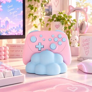 Pode incluir: Um comando de videojogos sem fios cor-de-rosa pastel com bot&otilde;es e joysticks azul-claros, pousado num suporte em forma de nuvem azul-clara. O comando apresenta uma disposi&ccedil;&atilde;o padr&atilde;o com um pad direcional, dois joysticks e v&aacute;rios bot&otilde;es.