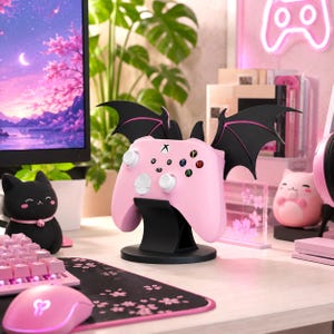 Könnte beinhalten: Ein rosafarbener Videospiel-Controller ruht auf einem schwarzen Ständer mit großen, schwarzen fledermausartigen Flügeln. Der Controller verfügt über weiße Analogsticks, ein weißes Steuerkreuz und farbige Aktionstasten. Der Ständer hat einen runden Fuß.
