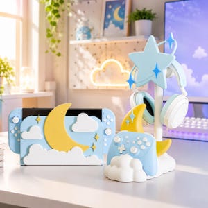 May include: A light blue gaming console dock and a matching controller stand featuring a yellow crescent moon, white clouds, and star accents. The console dock holds a handheld device, while the controller stand supports a wireless controller and a pair of light blue over-ear headphones with a large star topper.
