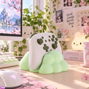 Pode incluir: Um comando de videojogos branco com bot&otilde;es e joysticks verde-azeitona escuro, pousado num suporte em forma de nuvem verde-menta claro. O comando apresenta uma disposi&ccedil;&atilde;o padr&atilde;o com um direcional, bot&otilde;es de a&ccedil;&atilde;o e dois joysticks anal&oacute;gicos.