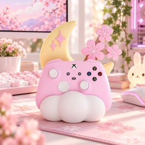 Peut inclure: Un support de manette Xbox rose avec un croissant de lune jaune orné de deux étoiles roses, trois fleurs de cerisier roses et une base en forme de nuage blanc. Le socle comprend des découpes pour les boutons et le logo Xbox central.