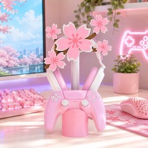 Könnte beinhalten: Ein rosafarbener Gaming-Zubehörständer mit einem zentralen Pfosten, der von fünf hellrosa Kirschblüten gekrönt wird. Der Ständer hält einen weiß-rosa Kopfhörer an den oberen Armen und einen rosa Videospiel-Controller auf der Basis.