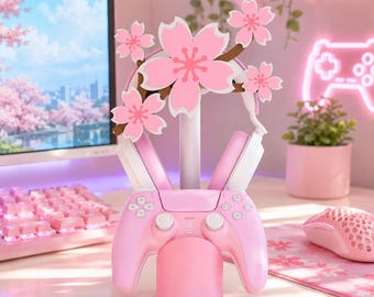 Pode incluir: Um suporte para acessórios de jogos rosa com um poste central coroado por cinco flores de cerejeira rosa claro. O suporte segura um par de auscultadores brancos e rosa nos braços superiores e um comando de videojogos rosa na base.