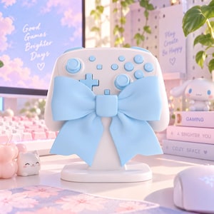 Puede incluir: Un soporte blanco para mando de videojuegos con un gran lazo decorativo de color azul claro en la parte frontal. El soporte tiene un acabado mate suave y una base ancha y estable diseñada para sostener el mando en posición vertical.