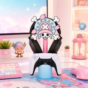 May include: A light blue and pink headphone and controller stand featuring a large circular emblem with a skull, crossbones, and cherry blossom petals. The stand holds a pair of black over-ear headphones and a white and black gaming controller.