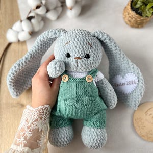 Puede incluir: Un peluche de conejito tejido de color azul claro con orejas largas y un peto verde. El conejito tiene una camisa blanca y botones de madera. La oreja tiene un parche blanco en forma de corazón con el texto "Easter 2026".