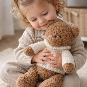 Könnte beinhalten: Ein brauner Teddybär mit einem weißen Pullover wird von einem kleinen Kind gehalten. Der Bär hat ein strukturiertes, flauschiges Aussehen. Das Kind trägt ein helles Outfit und sitzt auf einem weißen Teppich.