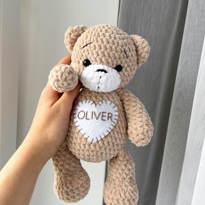 Könnte beinhalten: Ein gehäkelter beigefarbener Teddybär aus weichem Plüschgarn. Der Bär hat eine weiße Schnauze, schwarze Sicherheitsaugen und einen weißen Filzherzaufnäher auf der Brust, auf dem der Name OLIVER in braunem Garn mit sichtbaren weißen Stichen gestickt ist.