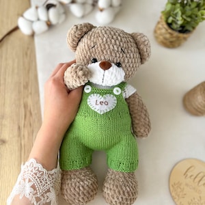 Puede incluir: Un oso de peluche hecho de hilo marrón suave con un hocico blanco y una nariz marrón. El oso lleva un peto de punto verde con un parche blanco en forma de corazón en el pecho con el nombre Leo bordado en hilo marrón y dos botones blancos.