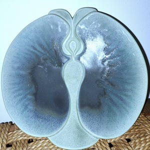 May include: A ceramic dish with a symmetrical, organic shape resembling a stylized butterfly or leaf. The surface features a reactive glaze with a matte light green border transitioning into a deep, dark gray center with speckled textures.