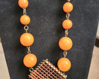 May include: A necklace featuring a series of smooth, round, bright orange beads connected by bronze-toned metal links. The central pendant consists of a rectangular grid structure with an amber-colored cabochon set in a circular filigree frame.