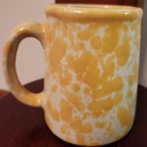 May include: A ceramic mug featuring a yellow and white sponge-painted or speckled pattern. The mug has a cylindrical body with a thick, rounded rim and a curved handle on the side. The surface has a glossy finish with irregular yellow spots.
