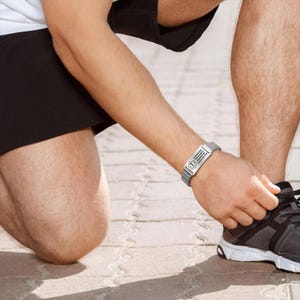 May include: A silver medical ID bracelet with a mesh band is worn on a person's wrist. The rectangular plate displays a medical symbol and text. The person is tying their shoe, wearing black shorts and gray athletic shoes.