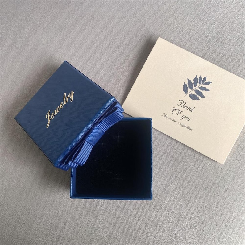 May include: A navy blue jewelry box with a gold "Jewelry" inscription, a blue ribbon, and a black velvet interior. A cream-colored card with a blue leaf design and the words "Thank You" is next to the box.