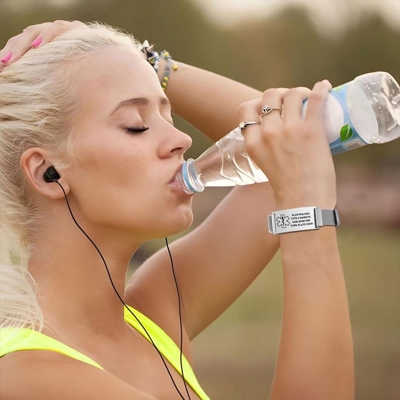 May include: A woman drinking water from a clear plastic bottle, wearing a silver medical ID bracelet. The bracelet has a black mesh band and white text. She is wearing a yellow top and black earbuds.