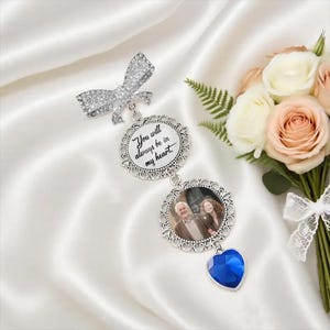 Könnte beinhalten: Ein silberfarbenes Foto-Medaillon mit einer schleifenförmigen Nadel, zwei kreisförmigen Rahmen und einem blauen herzförmigen Anhänger. Der obere Rahmen trägt den Text "You will always be in my heart". Der untere Rahmen enthält ein Foto. Ein Rosenstrauß ist im Hintergrund.