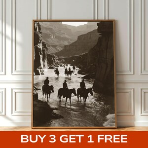 May include: A framed sepia-toned wall art print depicting a group of riders on horseback crossing a shallow river through a deep canyon. The scene features rocky cliffs on both sides and silhouettes of people in cowboy hats riding through water.