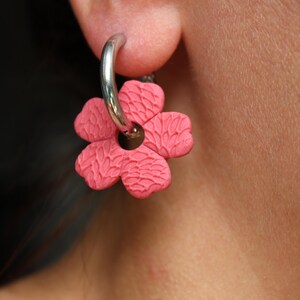 Peut inclure: Une boucle d'oreille créole en métal argenté avec une breloque en forme de fleur rose mat. La fleur présente une texture en écailles et une découpe circulaire centrale, suspendue à l'anneau en métal argenté lisse et poli.