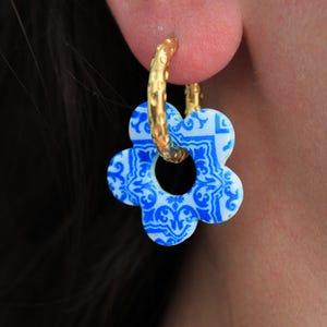 Puede incluir: Un pendiente de aro dorado con superficie texturizada y un dije colgante en forma de flor. El dije es blanco con un intrincado patrón floral azul que recuerda a los diseños de azulejos tradicionales, suspendido del aro circular dorado.