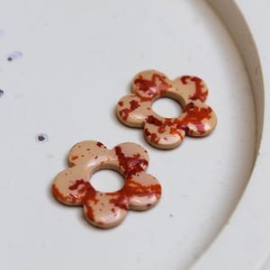 Könnte beinhalten: Zwei blütenförmige Dekorationsgegenstände mit beigefarbener Basis und rot-orangen Sprenkelmustern. Die Objekte haben ein Loch in der Mitte und sind auf einer weißen Oberfläche platziert. Es handelt sich wahrscheinlich um Ohrringe oder dekorative Elemente.