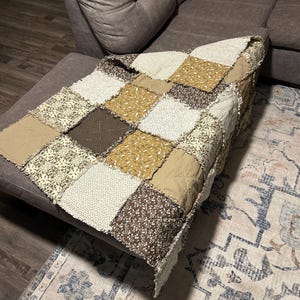 May include: A square patchwork quilt featuring a variety of fabric squares in shades of brown, tan, cream, and mustard yellow. The quilt is constructed with exposed, frayed seams between each square, creating a textured, grid-like appearance.